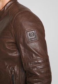 Hnědá kožená bunda s kapsou na zip, prošívanými detaily na ramenou a texturovanou nášivkou s logem na rukávu.