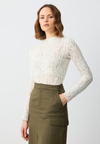 Haut en dentelle blanche à manches longues avec motif floral, associé à une jupe verte dotée de poches avant et d'une coupe droite.