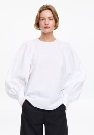 Personne aux cheveux courts châtain clair porte un chemisier blanc à manches volumineuses et un pantalon noir, debout devant un fond blanc uni.