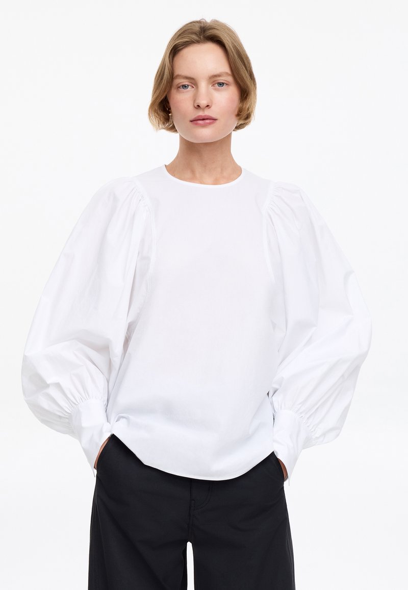 Personne aux cheveux courts châtain clair porte un chemisier blanc à manches volumineuses et un pantalon noir, debout devant un fond blanc uni.
