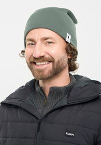 Smiling man with beard wearing green beanie, black quilted jacket, and gray zip-up sweater against white background.