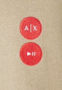Tessuto beige con due bottoni ricamati rossi. Il bottone superiore presenta il testo "A/X", mentre il bottone inferiore ha un'icona di riproduzione con linee di pausa.