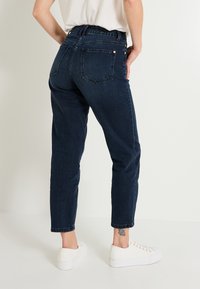 Persona con jeans azul oscuro y zapatillas blancas, vista desde atrás, de pie contra un fondo blanco liso.