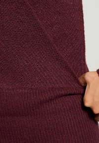 Nahaufnahme einer Hand, die einen strukturierten, gerippten, dunkelburgunderfarbenen Strickstoff eines Pullovers gegen einen schlichten hellen Hintergrund zieht.