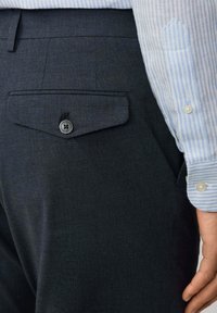 Närbild av mörka texturerade byxor med en knäppt bakficka och en del av en ljusblå randig skjortärm och hand synlig.