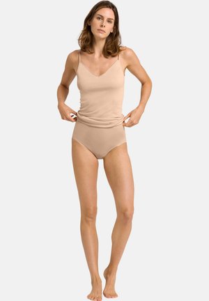 Vrouw draagt een beige hemdje en bijpassend high-waist ondergoed, waarbij ze de zoom van het hemdje aanpast, staand op blote voeten tegen een witte achtergrond.