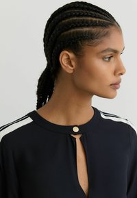 Sieviete ar tumšām pītām matu saknēm, ģērbusies melnā topā ar baltām svītrām uz pleciem un apaļu izgriezumu ar zelta pogu.