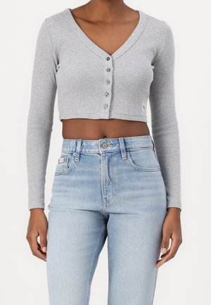 Mujer vistiendo un cárdigan corto gris de manga larga con textura acanalada y botones, combinado con jeans de tiro alto en color azul claro.