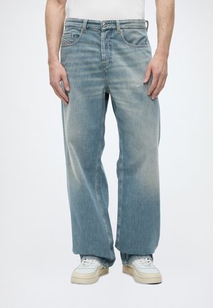 Homme portant un jean bleu clair ample et des baskets blanches, debout devant un fond clair uni.