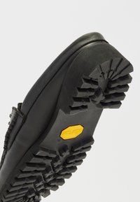 Chaussure en cuir noir avec une semelle en caoutchouc Vibram présentant un motif de crampons, conçue pour l'adhérence et la durabilité. Texture lisse et forme arrondie.