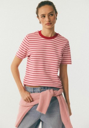 Kobieta w czerwono-biało paskowanej koszulce i jasnoniebieskich dżinsach z różowym swetrem związanym w pasie, patrząca w bok.