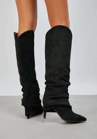 Bottes en suede noires au-dessus du genou avec un design flou, une pointe effilée et un petit talon bloc, présentant une texture lisse et un style minimaliste.