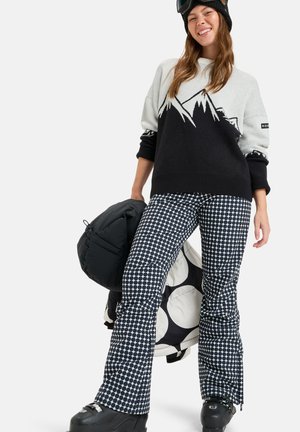 Sweater in light gray and black with mountain graphic, paired with black and white polka dot snow pants. Holding a black puffy bag, wearing ski boots.