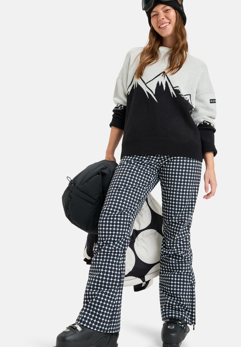 Sweater in light gray and black with mountain graphic, paired with black and white polka dot snow pants. Holding a black puffy bag, wearing ski boots.