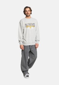 Sudadera gris con gráfico "DC Shoes Worldwide" en azul y amarillo. Combinada con jeans grises holgados y zapatillas negras y blancas.