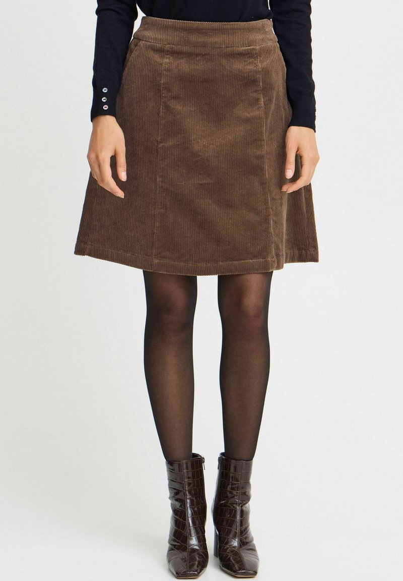 Jupe évasée en velours côtelé marron avec des nervures horizontales, une taille et un ourlet détaillés. Associée à des collants noirs et des bottes marron en cuir texturé crocodile.