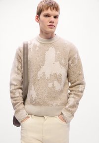 Young man wearing a beige and cream textured sweater with ribbed cuffs and hem, cream pants, and a taupe shoulder bag strap.