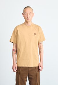 Tan cotton t-shirt with a crew neck and short sleeves. Features a small heart-shaped logo on the left chest. Smooth texture.