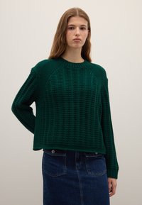 Grüner Strickpullover mit Rippenstruktur und Raglanärmeln. Grob gestricktes Garn und Rundhalsausschnitt. Kombiniert mit einem Jeansrock.
