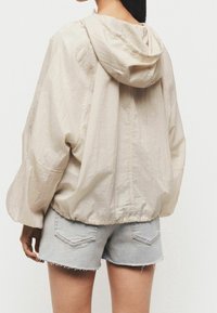 Veste beige légère et courte avec capuche, en tissu texturé et ourlet élastique. Assortie à un short en jean bleu clair effiloché.