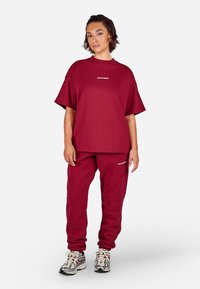Bordó pamut póló és melegítőnadrág szett laza szabással. A póló környakú és nyomott logóval díszített; a nadrág alja mandzsettás.