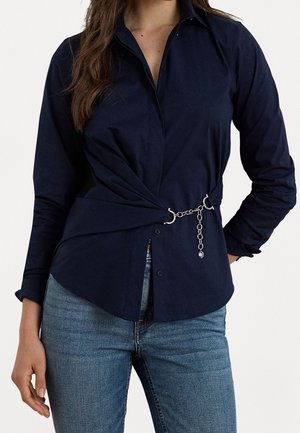 Vrouw draagt een marineblauw shirt met lange mouwen, een gedraaide voorkant en een zilveren ketting, gecombineerd met blauwe spijkerbroeken.