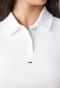 Närbild av en person som bär en vit ribbad polo t-shirt med en liten röd, vit och blå rektangulär logotyp på bröstet.