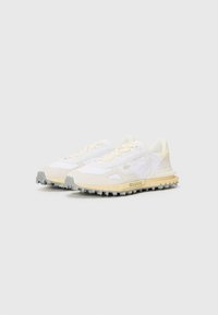 Witte hardloopschoenen met een gestructureerde stof en een bovenkant van suede, met gele accenten, een lichtgrijze zool en een robuuste buitenzool met spikes.