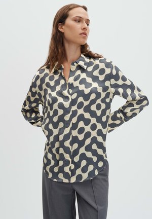 Long-sleeve button-up shirt in beige with a dark wave pattern; smooth fabric, classic collar, and curved hemline. Paired with gray trousers.