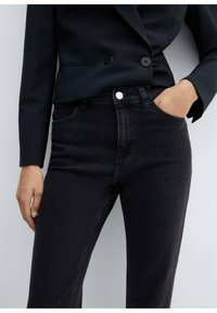 Jeans de mezclilla negra con cintura alta, bolsillos frontales y cierre de botón plateado. Combinados con un blazer negro ajustado. Textura suave.