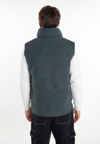Dunkelgrüne ärmellose Weste mit hohem Kragen, mit einer gesteppten Textur und horizontalen Nahtlinien, getragen über einem hellen Pullover.