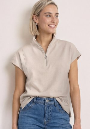Lyshåret kvinde smiler, iført beige kortærmet top med frontlynlås og blå jeans mod en ensfarvet lys baggrund.