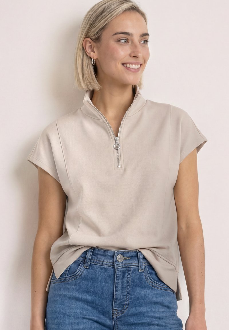 Femme blonde souriante, portant un haut beige à manches courtes avec fermeture éclair devant et un jean bleu sur un fond clair uni.