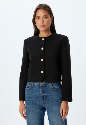 Zwarte cropped jacket van textiel met een ronde halslijn en zes gouden knopen aan de voorkant. Gemaakt met blauwe jeans.