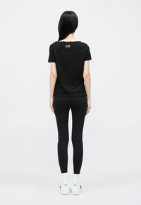 Fekete rövid ujjú póló EAZ logóval a hátulján, fekete leggingszel és fehér tornacipővel, hátulnézetben viselve.