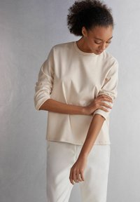 Beige Langarmoberteil mit entspanntem Schnitt und Rundhalsausschnitt. Weicher Stoff mit umgeschlagenen Bündchen, kombiniert mit hellen, körperbetonten Hosen.