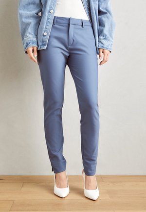 Person trägt eng anliegende blaue Hose, weiße High Heels, weißes Oberteil und eine hellblaue Jeansjacke, steht auf einem Holzboden.