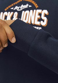 Karinis džemperis su įsiūtais rankogaliais. Artimas vaizdas rodo tekstūrą ir siuvimo detales, o fone matomas oranžinis logotipas.