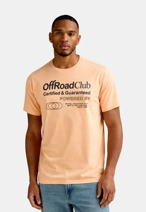 Muž nosi broskyňové tričko s čiernym nápisom "OffRoad Club Certified & Guaranteed Powered By Petrol Industries Co.", stojaci na jednoliatom pozadí.