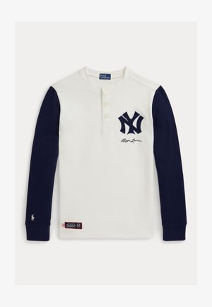 Ilgųjų rankovių henley marškiniai, pagaminti iš medvilnės, su balta kūno dalimi, tamsiai mėlynomis rankovėmis, NY logotipu ant krūtinės, mygtukais ant apykaklės ir audiniu etiketėje ant hem. Išvaizda.