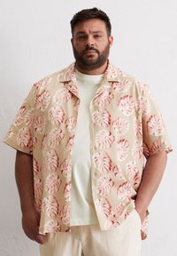 Korte mouwen shirt van beige katoen met een rood bladmuster. Heeft een notch-kraag en knopen. Draagt boven een lichtgekleurde T-shirt.