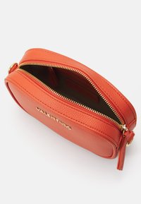 Borsa crossbody in pelle arancione con forma arrotondata, zip dorata e branding impresso sul davanti. La parte superiore aperta mostra l'interno.