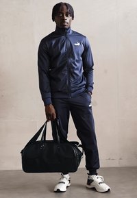 Marineblå træningsdragt med en lynlåsjakke og matchende bukser, parret med hvide og sorte sneakers. Holder en sort duffel bag med dobbelt håndtag.