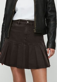 Gonna di denim marrone con design a pieghe e cuciture visibili. Presenta una chiusura frontale con bottoni e tasche laterali. Indossata con una giacca di pelle nera.