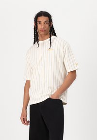 Hvid kortærmet t-shirt med lodrette gyldne striber og broderet logo på brystet, parret med sorte bukser. Løs pasform og glat stof.