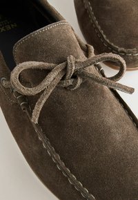 Mocassin en daim brun avec coutures apparentes et nœud de lacet attaché sur le dessus, montrant un gros plan des détails de la texture et de l'artisanat.