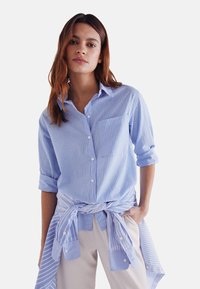 Light blue button-up shirt with white vertical stripes, long sleeves rolled up, and a chest pocket. Lower tied shirt shows matching fabric.