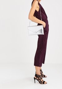 Metaliczna srebrna kopertówka o kształcie koperty, z łańcuszkiem i gładką fakturą, zestawiona z dopasowanym, głęboko fioletowym kombinezonem z falbanami oraz czarnymi sandałkami na obcasie.