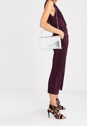 Clutch en métal argenté avec un design en enveloppe, une chaîne et une texture lisse, associé à une combinaison plissée ajustée de couleur violet foncé et des escarpins noirs à lanières.