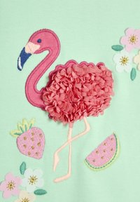 Izšūta piparmētru zaļa auduma virsma ar rozā flamingo, kam ir teksturētas spalvas, blakus atrodas rozā zemenes un ziedi ar zelta akcentiem.